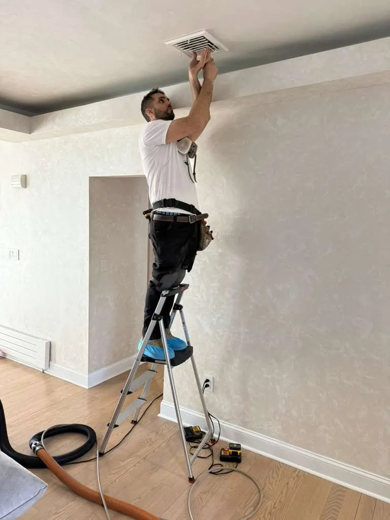 Air duct cleaning technician on ladder inspecting ceiling vent in Nassau County home